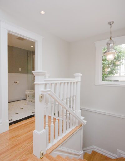 Interior view of a home featuring wooden floors, a white staircase, and a bathroom entrance with a glass door.